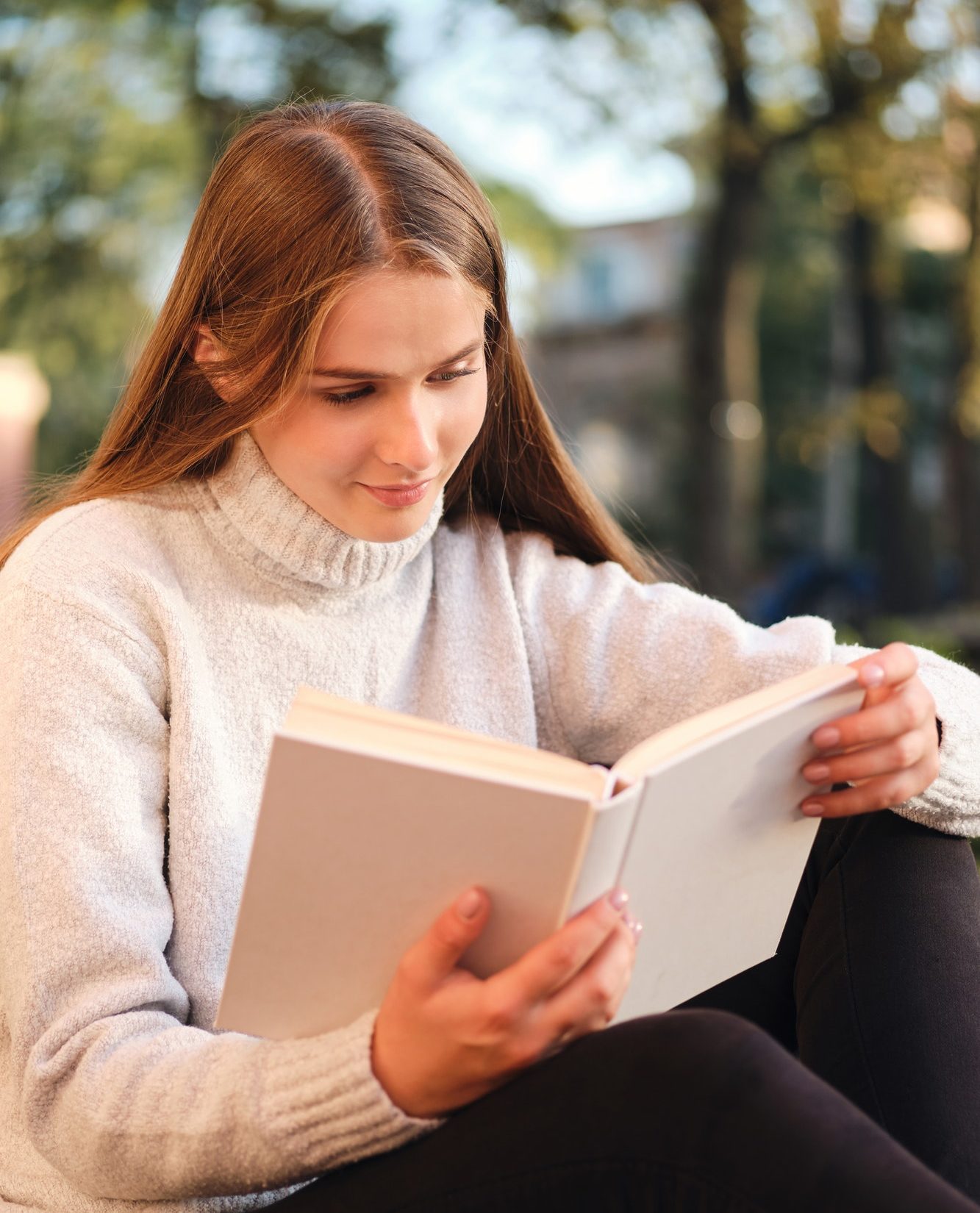 Young pretty student girl in cozy sweater dreamily reading book studying outdoor